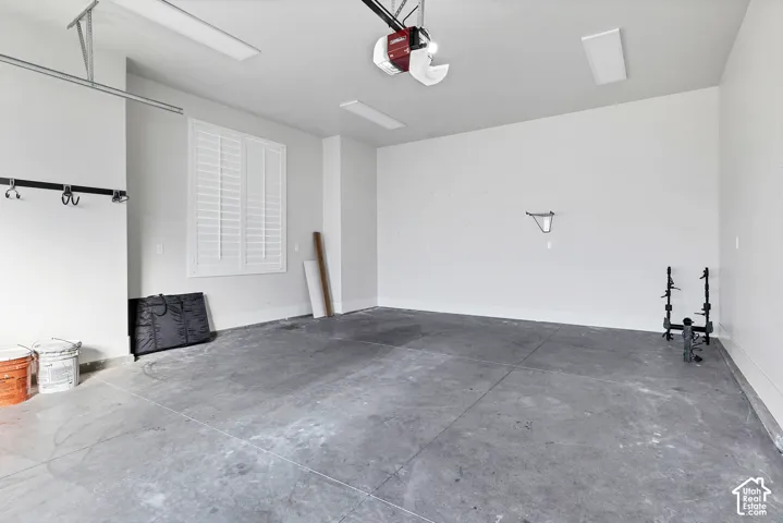 Concrete flooring, window with white plantation shutters, white walls, overhead garage door opener, overhead lighting