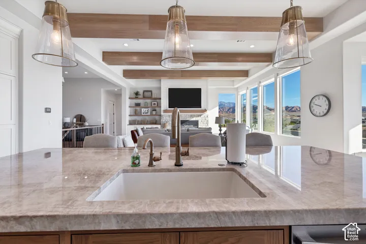Oversized kitchen island with a built-in sink, three pendant light fixtures, wood-finish ceiling beams, expansive windows, and a built-in fireplace with display shelving