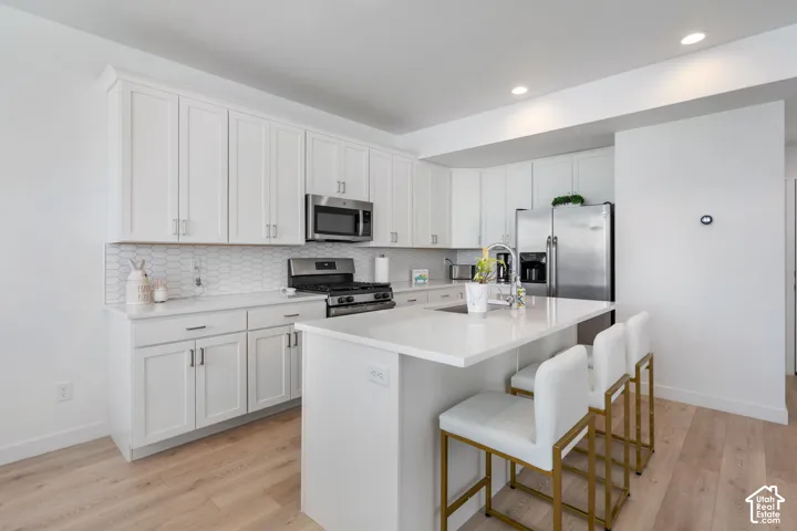 White shaker cabinetry, stainless steel appliances, central island with integrated sink, patterned tile backsplash, and wood-finish flooring