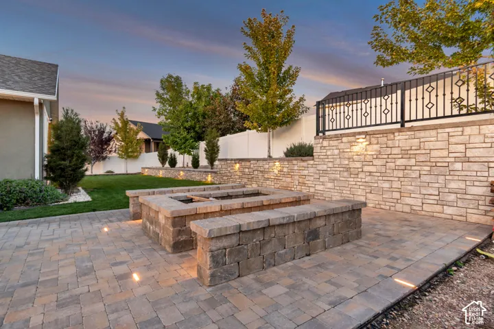 Paver patio with an integrated fire pit, stone-clad retaining wall with accent lighting, and decorative metal railing