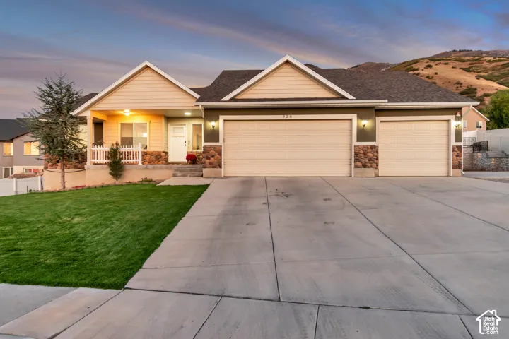 Expansive driveway, three-car garage, covered porch with white picket railing, stone accents on exterior, mountain backdrop