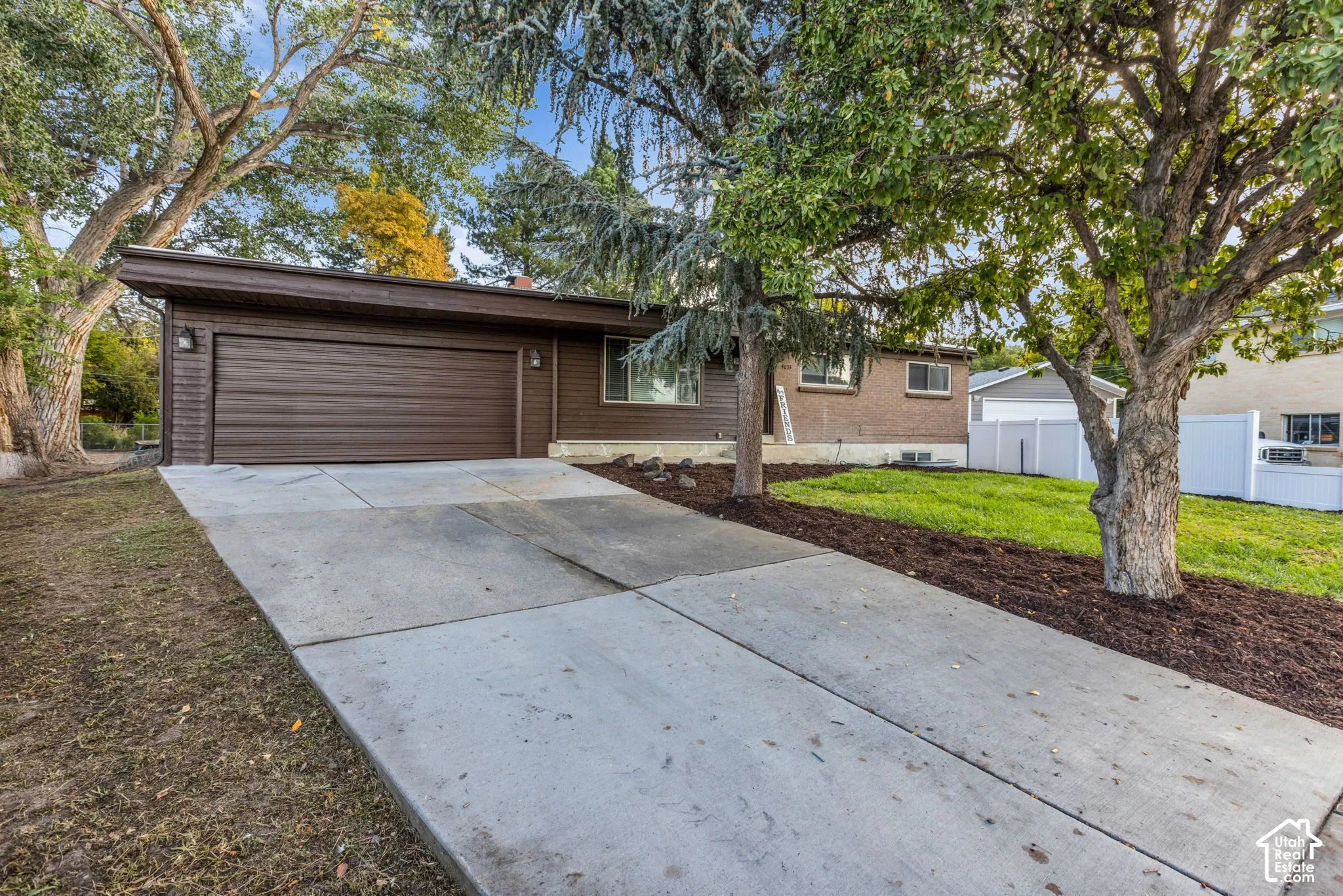 Brown horizontal siding, large attached garage, concrete driveway, mature trees, and landscaped yard with mulch