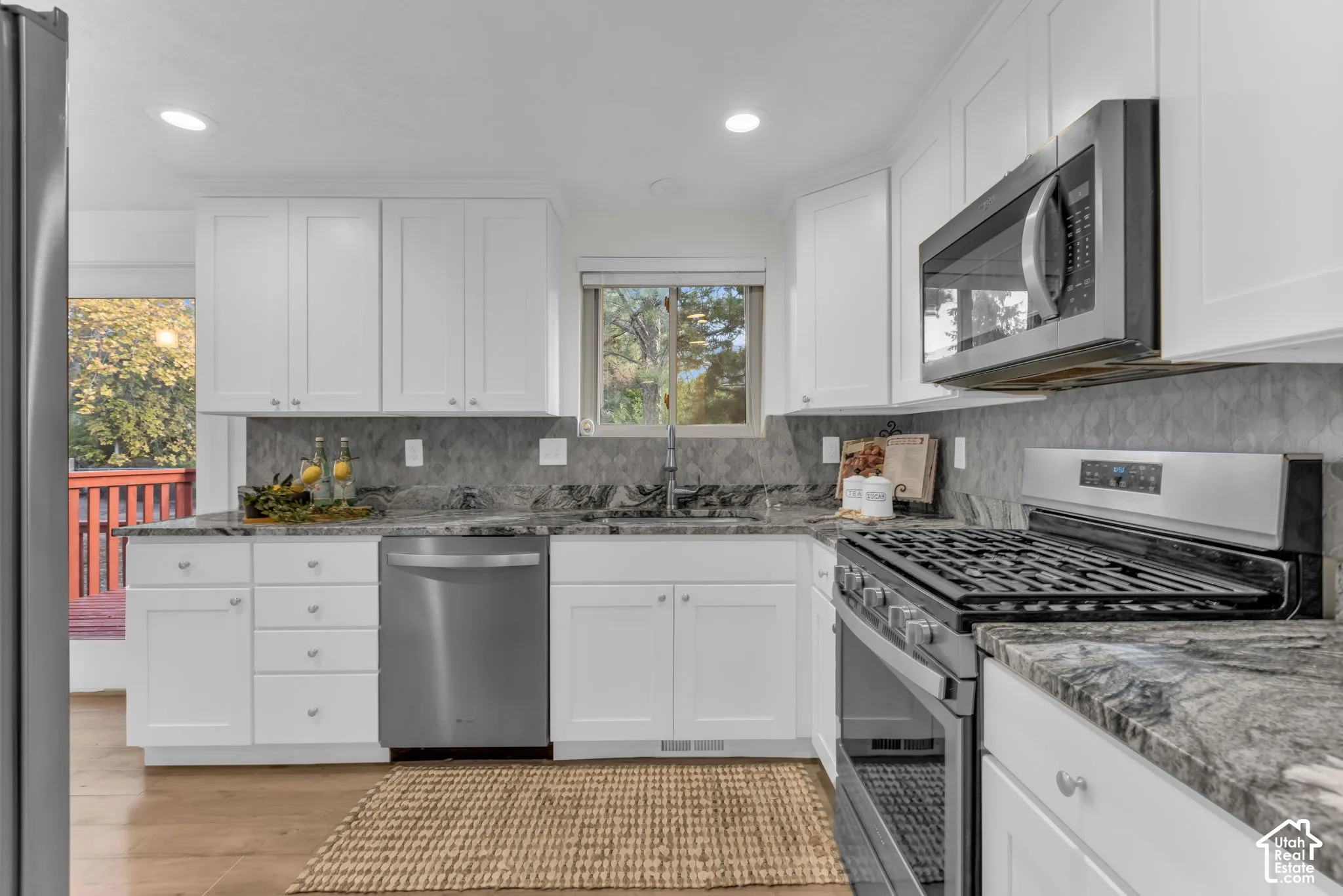 White shaker cabinetry with silver hardware, stone countertops with a gray tile backsplash, stainless steel appliances, and wood-finish flooring