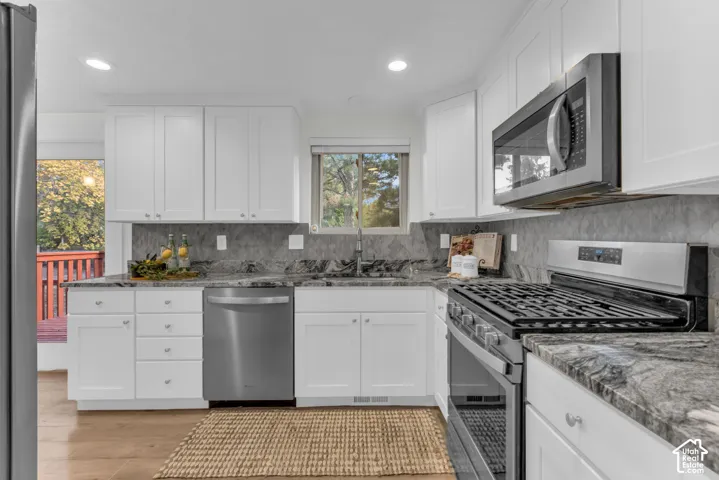 White shaker cabinetry with silver hardware, stone countertops with a gray tile backsplash, stainless steel appliances, and wood-finish flooring