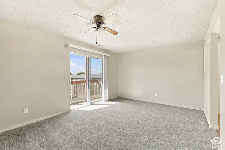 Spacious room with plush gray carpeting, a ceiling fan, and sliding glass doors leading to an exterior deck with wood railing
