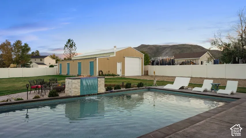 Expansive backyard featuring a rectangular swimming pool with integrated water feature, an adjacent paved patio, and a separate large utility building with multiple access doors and a garage door