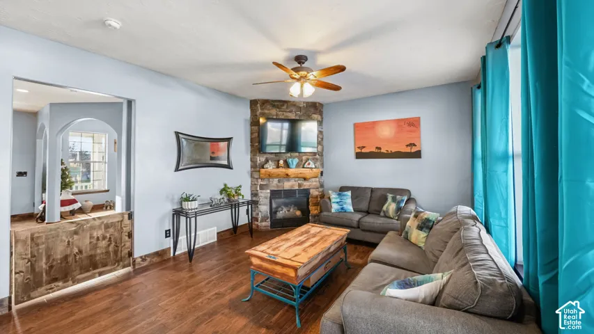 Wood-finish flooring, corner stone fireplace with wood mantel, ceiling fan, and arched doorway