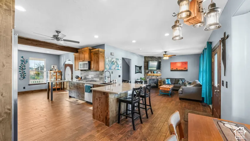 Open-concept living area featuring wood-finish flooring, a kitchen island with seating, natural wood cabinetry, a stone-clad fireplace, and recessed lighting