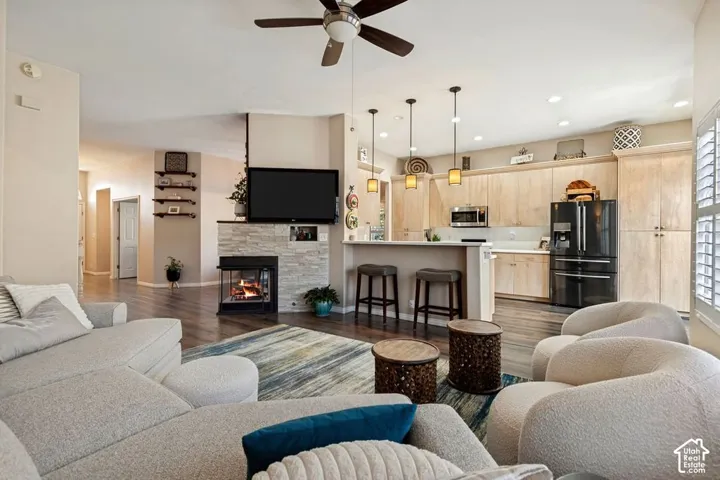 Open-concept living space featuring wood-finish flooring, a stone-faced fireplace, a kitchen island with pendant lighting, and light wood cabinetry