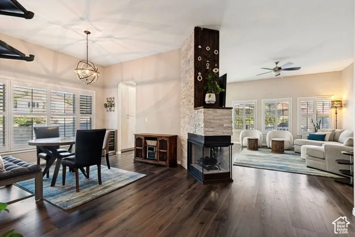 Rich wood-finish flooring, a two-sided fireplace with stone surround, white louvered window treatments, a ceiling fan, and an orb chandelier