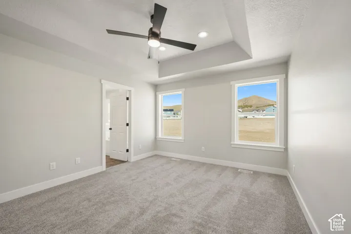 Tray ceiling with recessed lighting and a ceiling fan, two windows with white trim, and light gray carpet