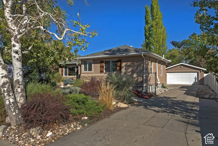 Stucco exterior with stone accent, front landscaping with mature trees, detached two-car garage, concrete driveway, and privacy fence