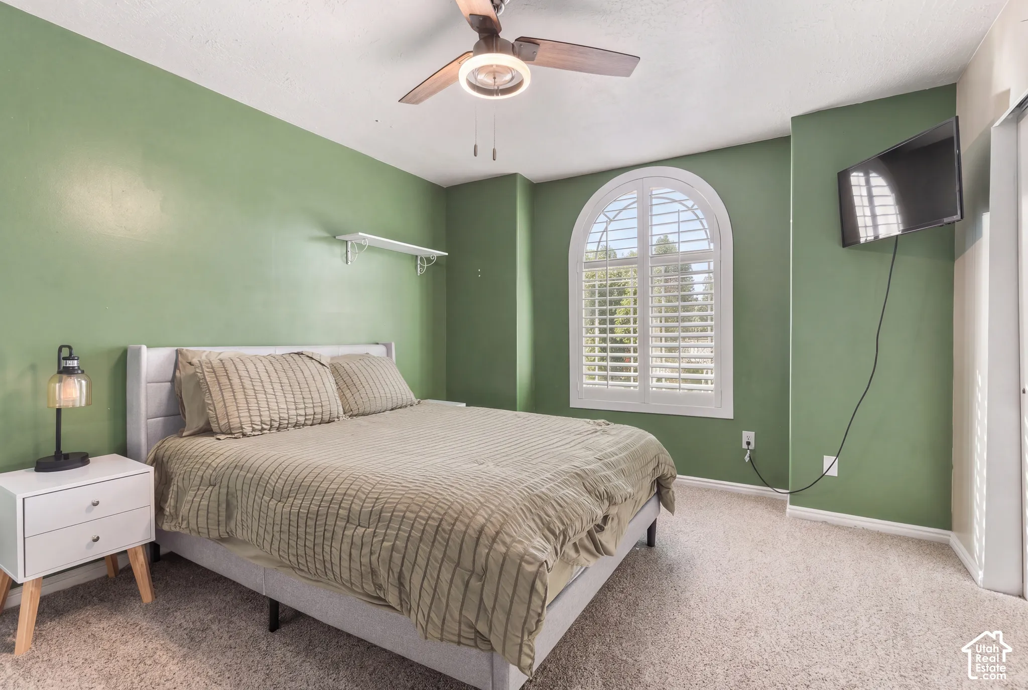 Arched window with plantation shutters, ceiling fan with integrated lighting, carpeted flooring, wall-mounted shelving