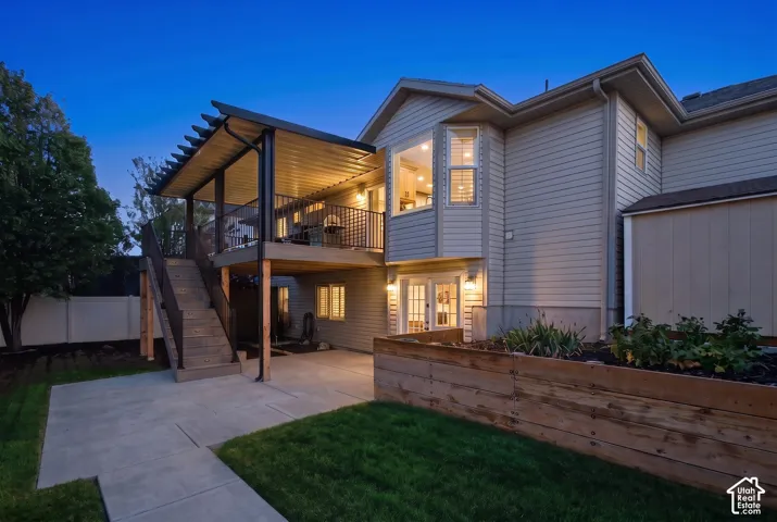 Covered elevated deck with black metal railing, exterior siding, ground-level concrete patio, bay window, and wood-finish retaining wall