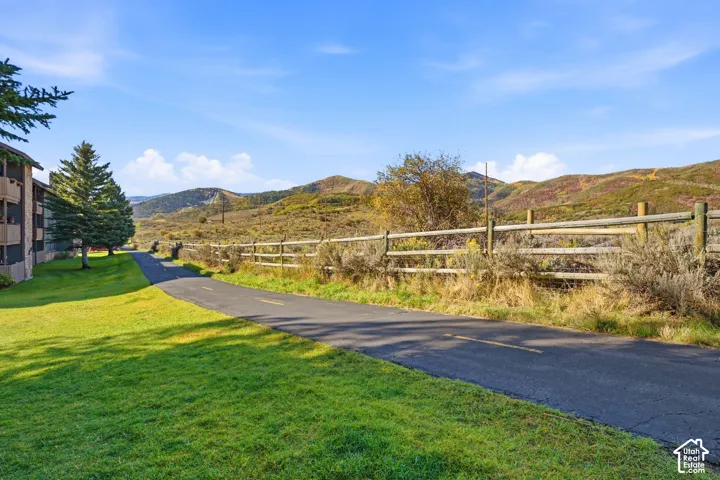 Paved pathway bordered by a split-rail fence, expansive grassy areas, and distant mountainous terrain