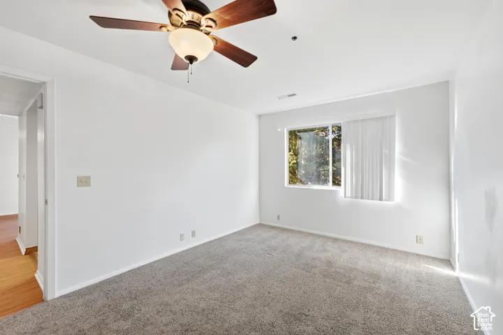 Neutral-toned carpeting, white walls, wood-finish ceiling fan with integrated lighting, and a single window with vertical blinds