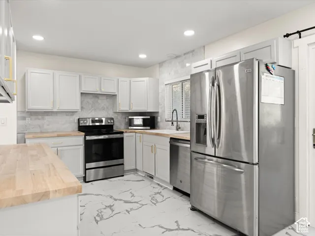 Kitchen featuring stainless steel appliances, light wood-finish countertops, white cabinetry with gold-tone hardware, a white tile backsplash, and white flooring with a gray veined pattern