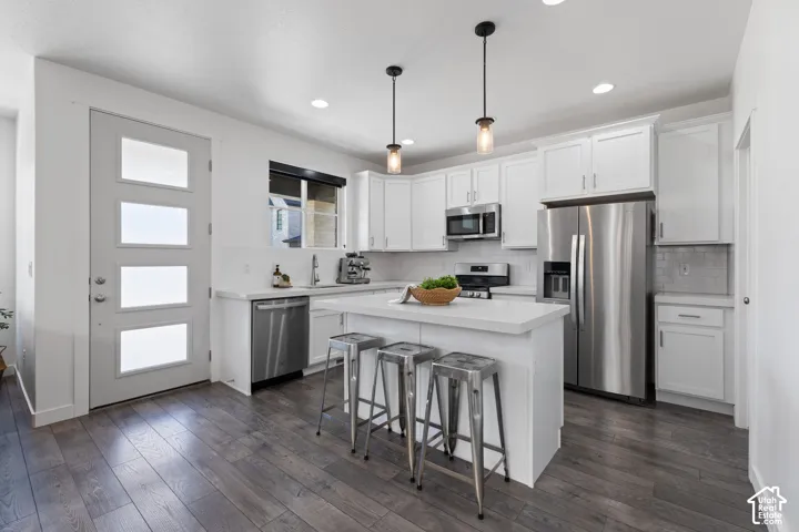 Modern kitchen featuring a central island, stainless steel appliances, white cabinetry, wood-finish flooring, and pendant lighting