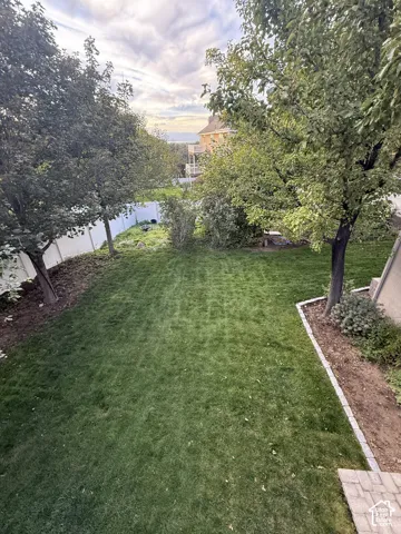 Expansive backyard featuring a manicured lawn, mature trees, white vinyl fencing, and a stone-edged garden bed