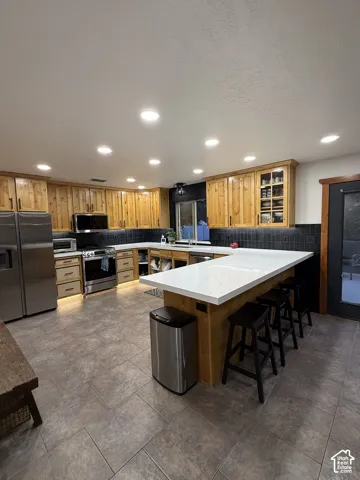 Recessed lighting illuminates a kitchen featuring wood cabinetry, stainless steel appliances, a white island countertop, and a dark tile backsplash