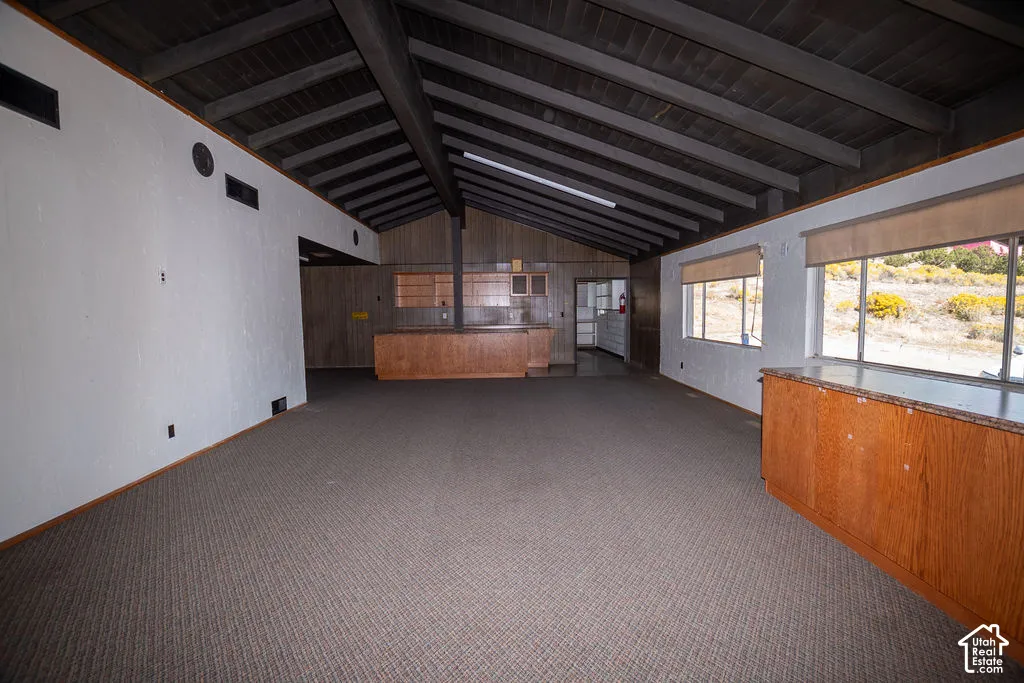 Vaulted ceiling with exposed wood beams, wood-paneled accent wall, built-in wood cabinetry with countertop, multiple windows with roller shades, and wall-to-wall carpeting