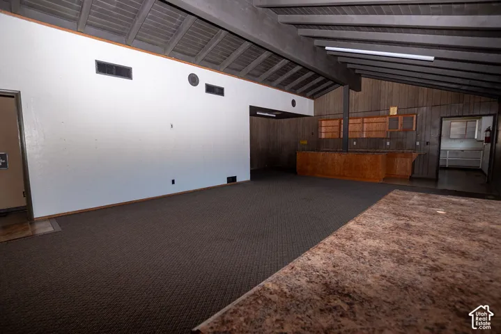 Vaulted ceilings with exposed beams, wood-paneled accent walls, built-in wooden bar, and textured carpet flooring