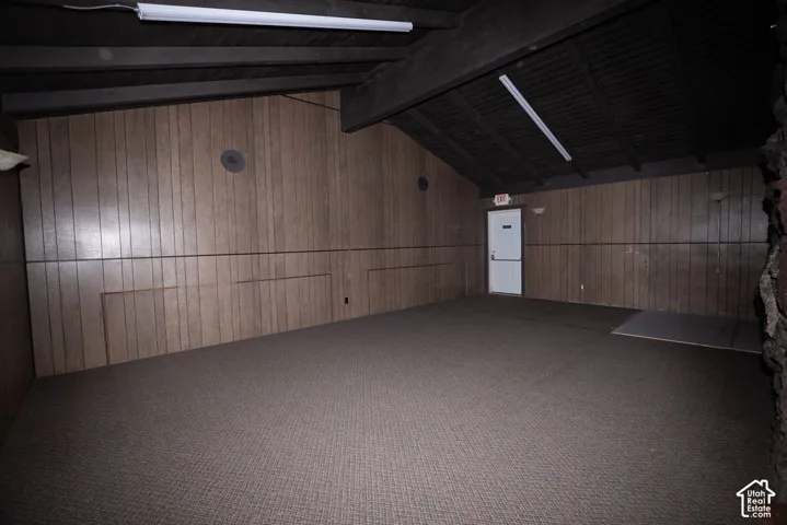 Vaulted ceiling with exposed beams, wood-paneled walls, wall-mounted sconces, and neutral carpet flooring