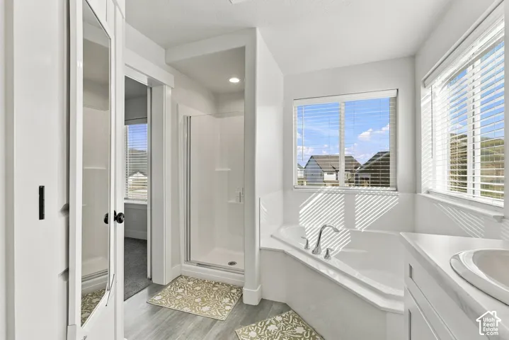 Corner soaking tub, two windows with blinds, built-in vanity with integrated sink, glass-enclosed shower, and wood-finish flooring