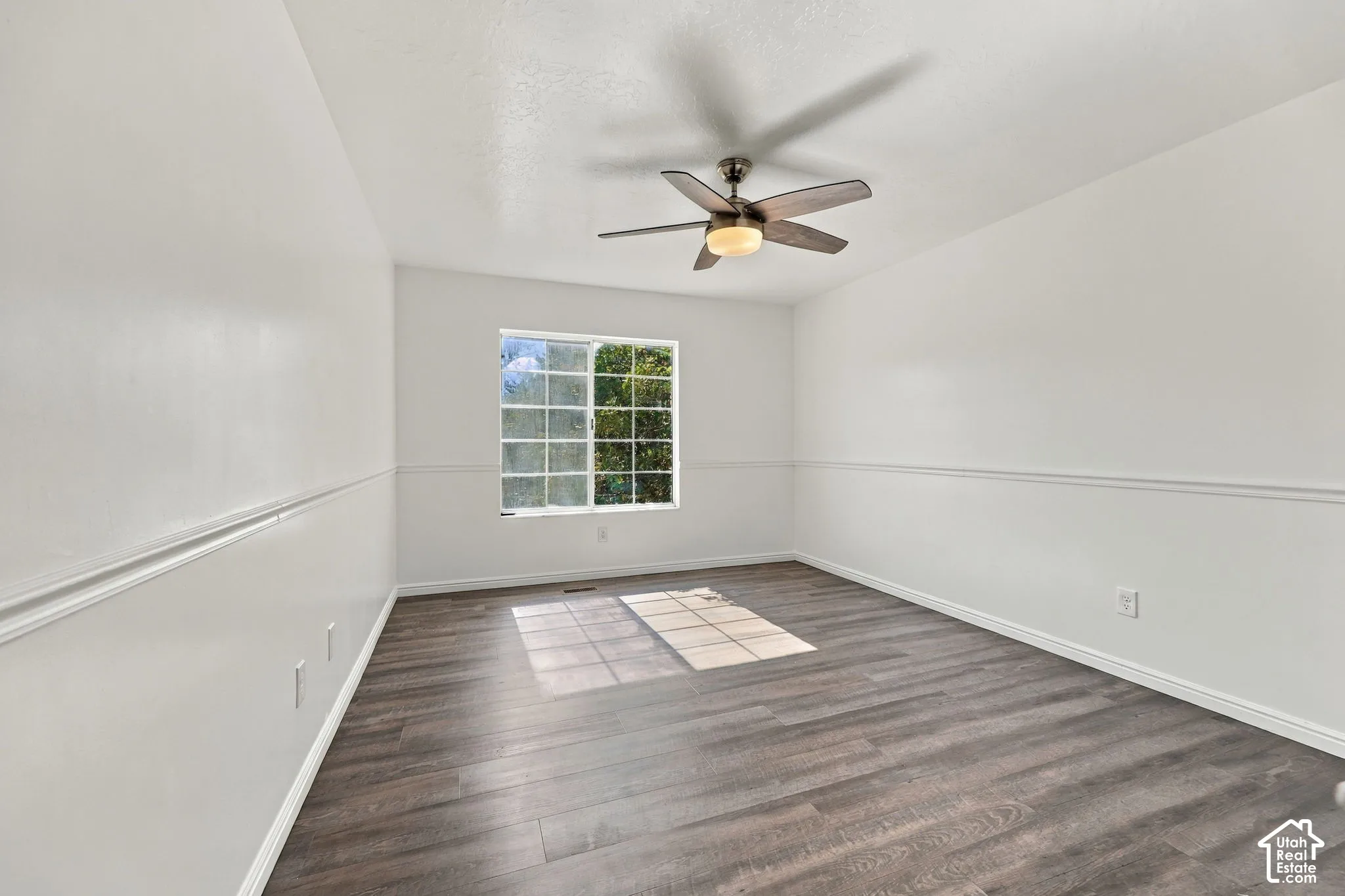 Wood-finish flooring, white walls with chair rail molding, ceiling fan with integrated lighting, and a multi-pane window