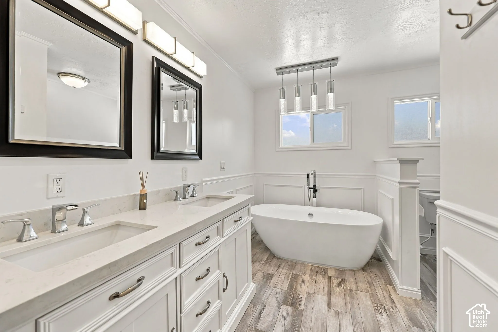 Double vanity with integrated sinks and chrome fixtures, standalone soaking tub, wood-finish flooring, two windows, and wainscoting
