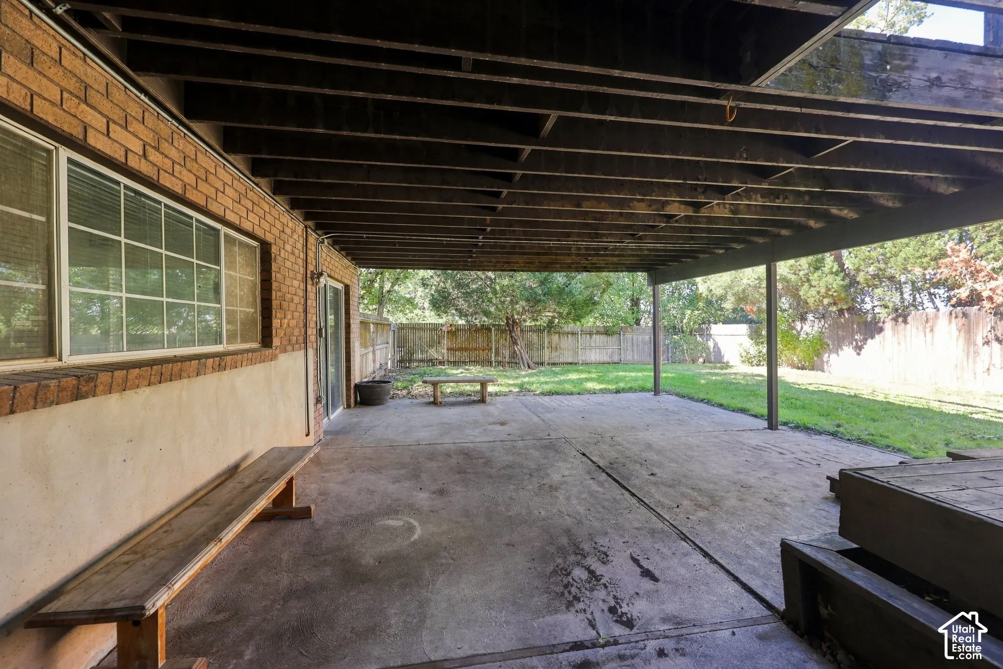 Covered concrete patio with exposed wood joists and support pillars, brick exterior wall, and sliding glass door