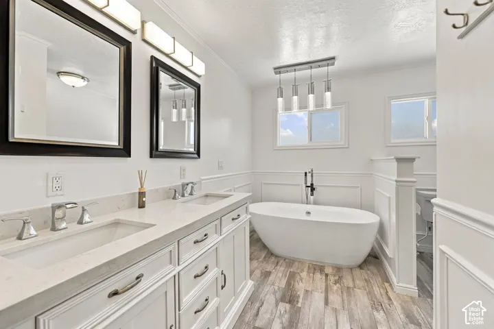 Double vanity with integrated sinks and chrome fixtures, standalone soaking tub, wood-finish flooring, two windows, and wainscoting