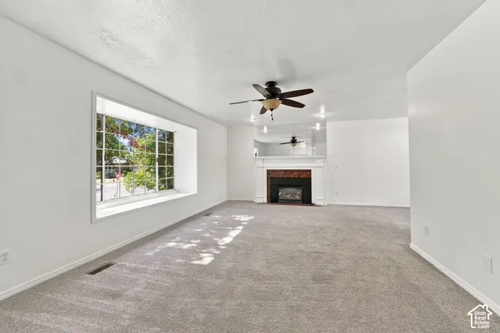 Expansive room featuring light gray carpeting, a window seat with a multi-pane window, a fireplace with a white mantel and brick surround, and a ceiling fan