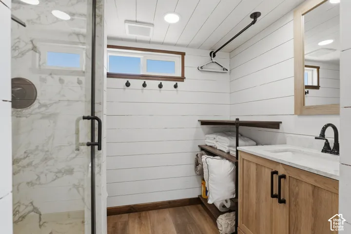 Wood-finish flooring with white shiplap walls, a wood-finish vanity with a white countertop and rectangular sink, a glass-enclosed shower with marble-look tile, and a wall-mounted drying rack