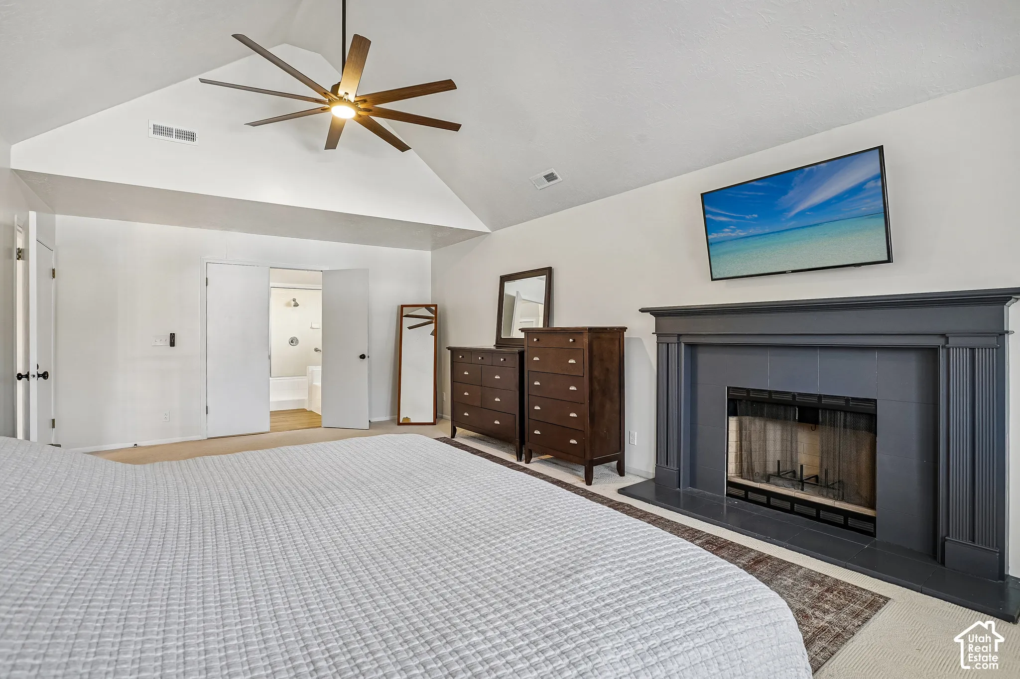 Vaulted ceiling with an overhead fan, carpet flooring, a dark-toned fireplace with a mantel, and double doors opening to a bathroom with a tub