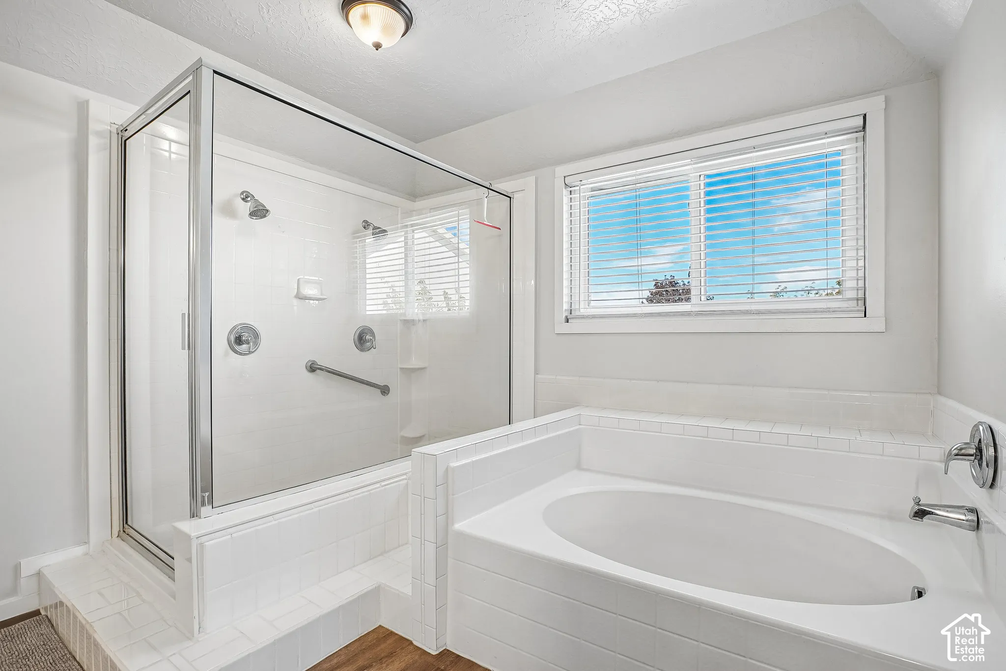White tiled soaking tub, separate glass-enclosed shower, chrome fixtures, and a window with blinds