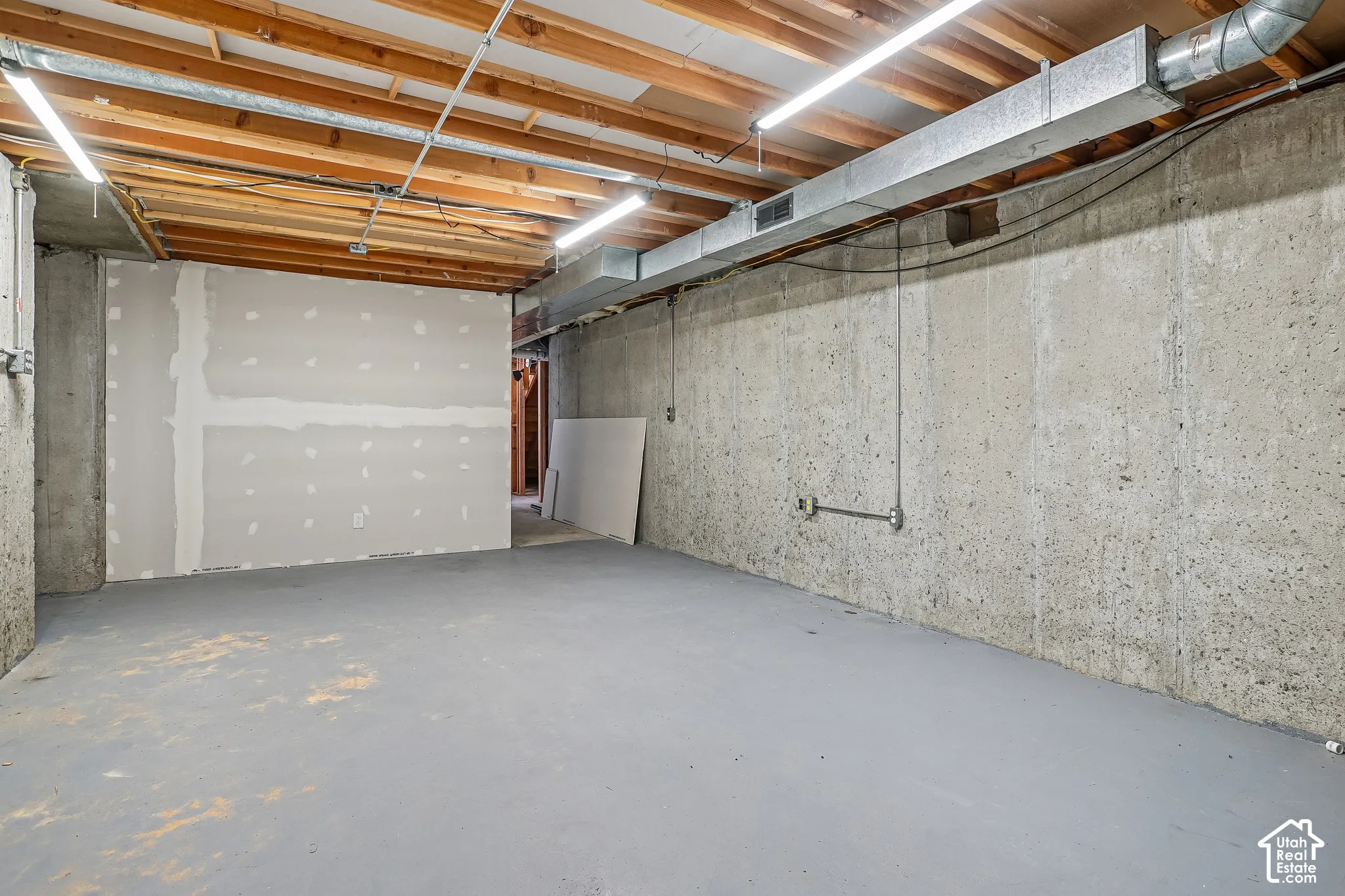 Unfinished basement space featuring exposed wooden ceiling joists, concrete walls, painted concrete flooring, and fluorescent lighting
