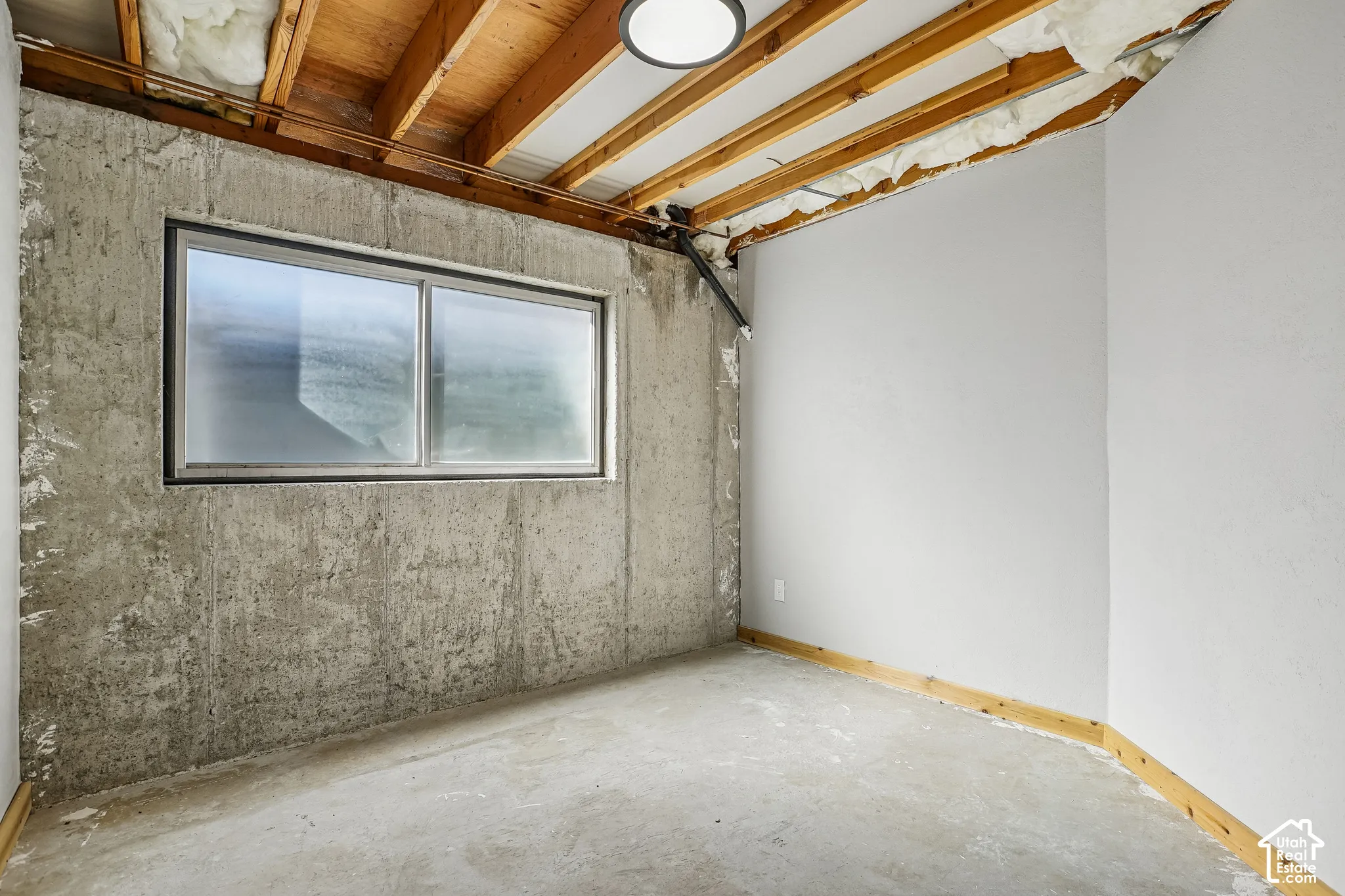 Concrete wall with a double-pane window, exposed wood ceiling joists, and a flush-mount overhead light fixture