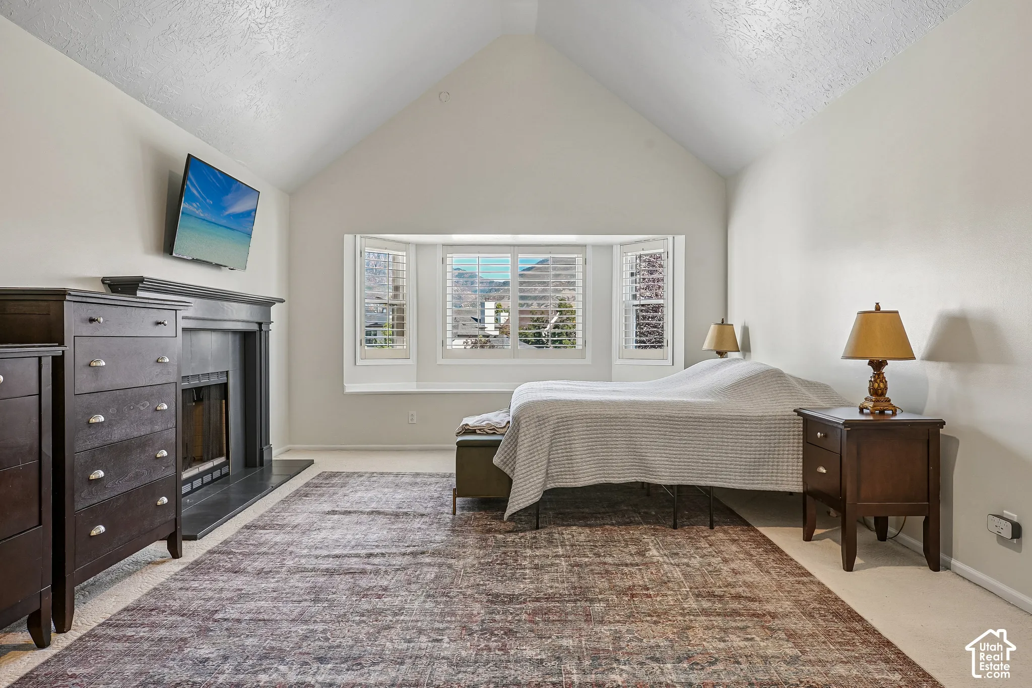 Vaulted ceiling with a bay window featuring white plantation shutters, and a fireplace with a dark mantel