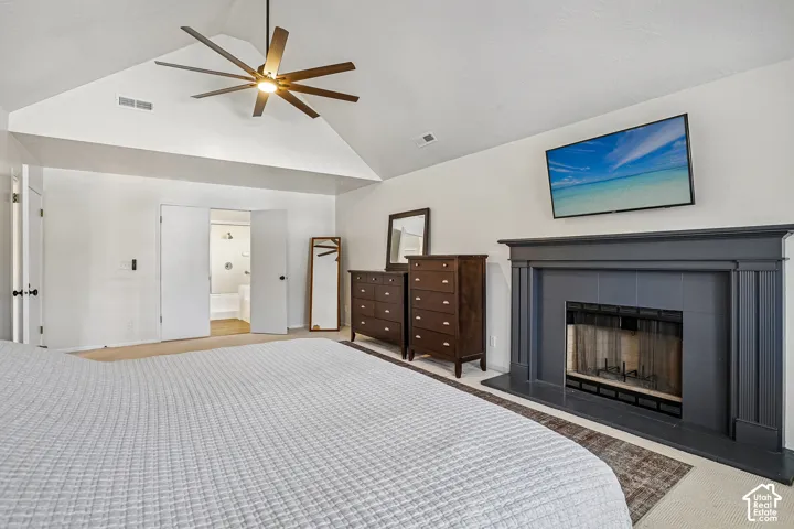 Vaulted ceiling with an overhead fan, carpet flooring, a dark-toned fireplace with a mantel, and double doors opening to a bathroom with a tub