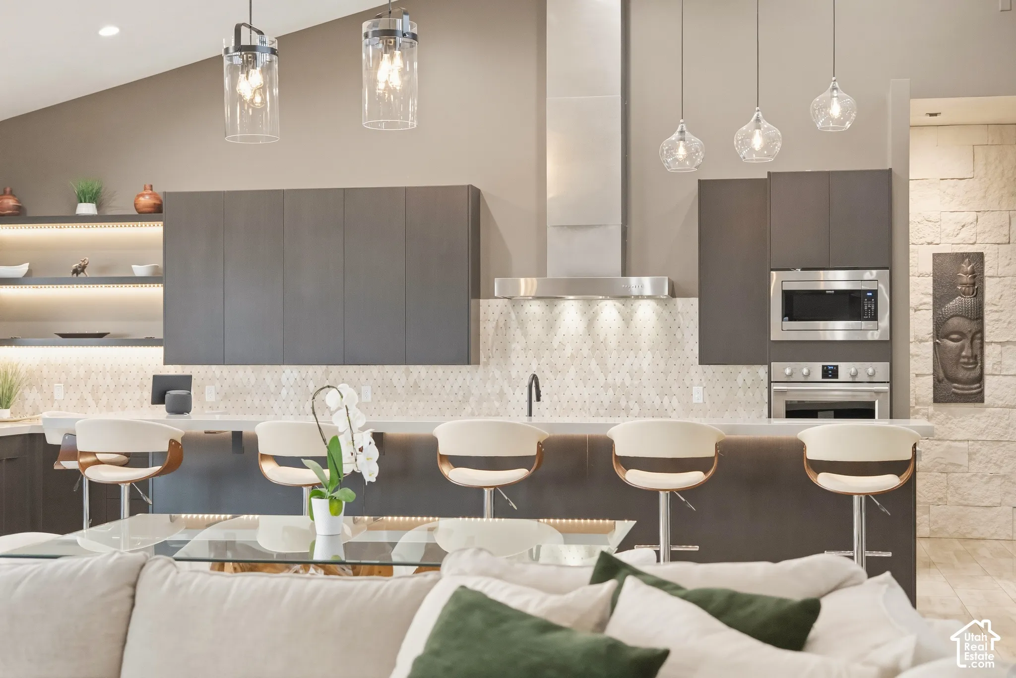 Vaulted ceiling with pendant lighting, integrated stainless steel appliances, modern cabinetry, built-in shelving with accent lighting, and a textured stone accent wall