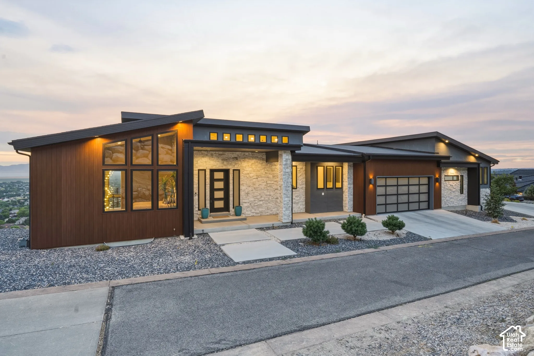 Modern architectural exterior featuring a sloped roofline, stacked stone accents, wood-finish siding, a frosted glass garage door, and a multi-pane entry door