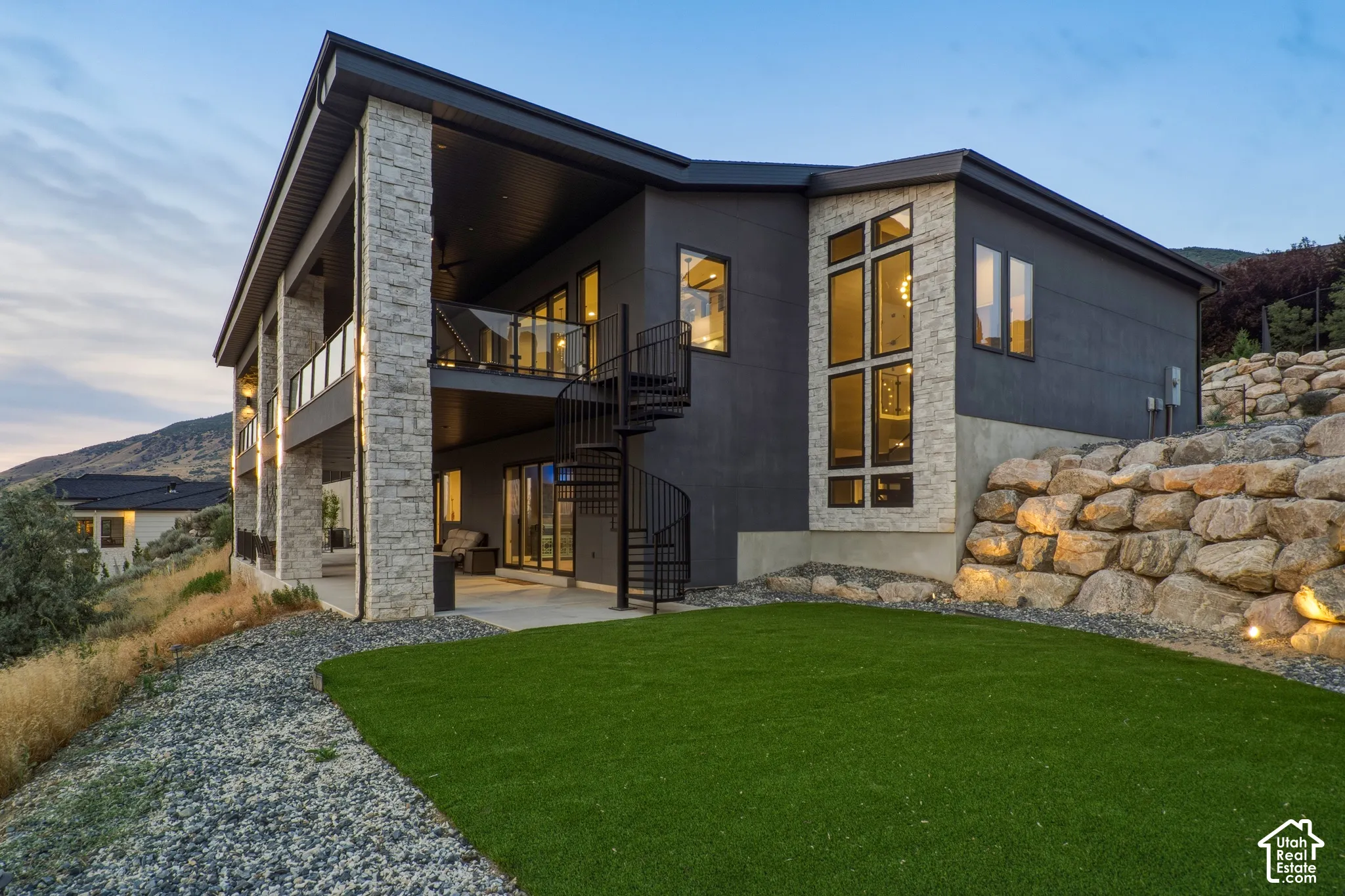 Modern exterior with dark siding and stone accents, featuring a multi-level deck with glass railings, a spiral staircase, and large picture windows