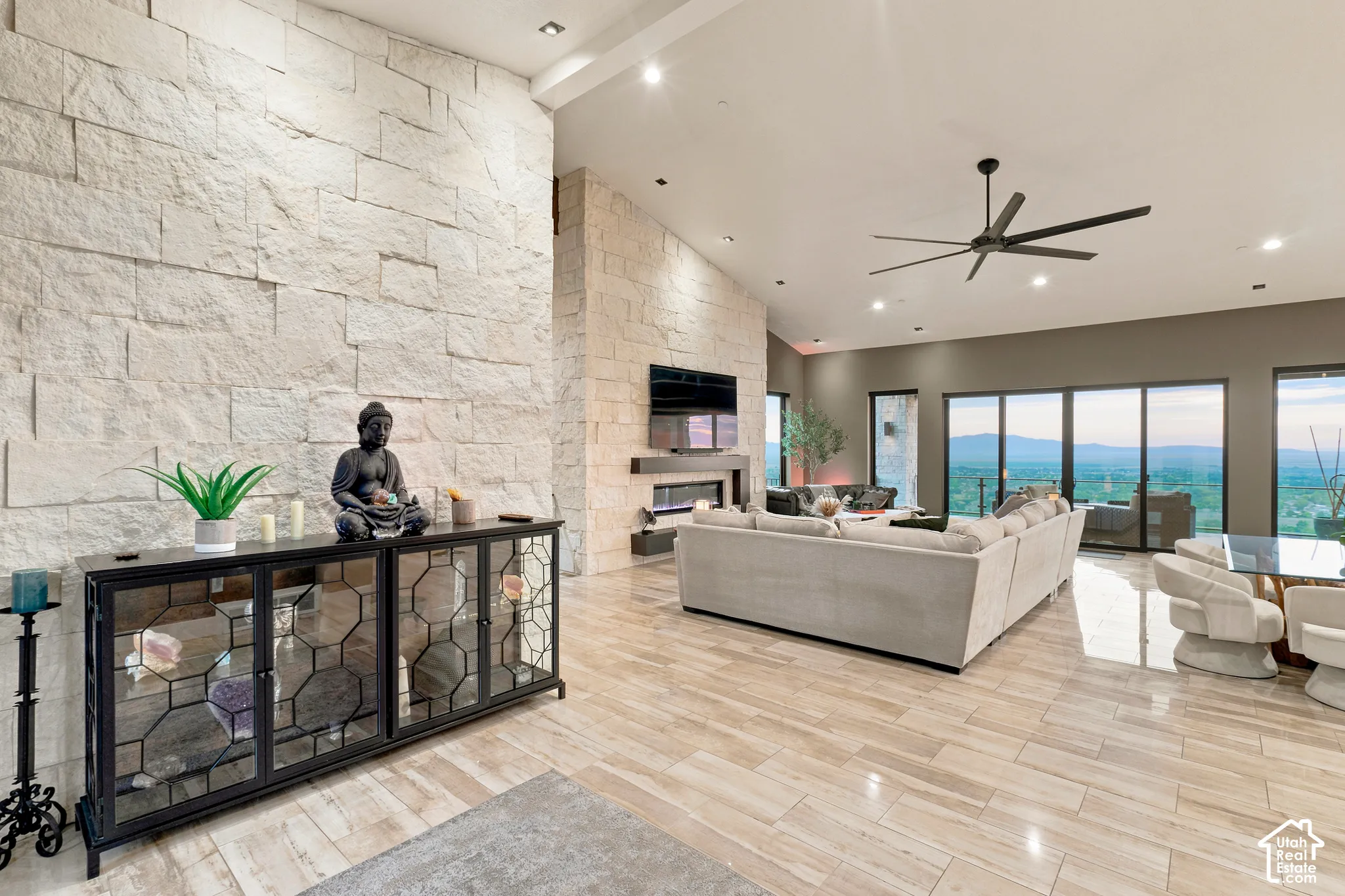 Vaulted ceiling with recessed lighting, floor-to-ceiling stone accent wall, fireplace with built-in media shelf, large format tile flooring, and multi-panel sliding glass doors