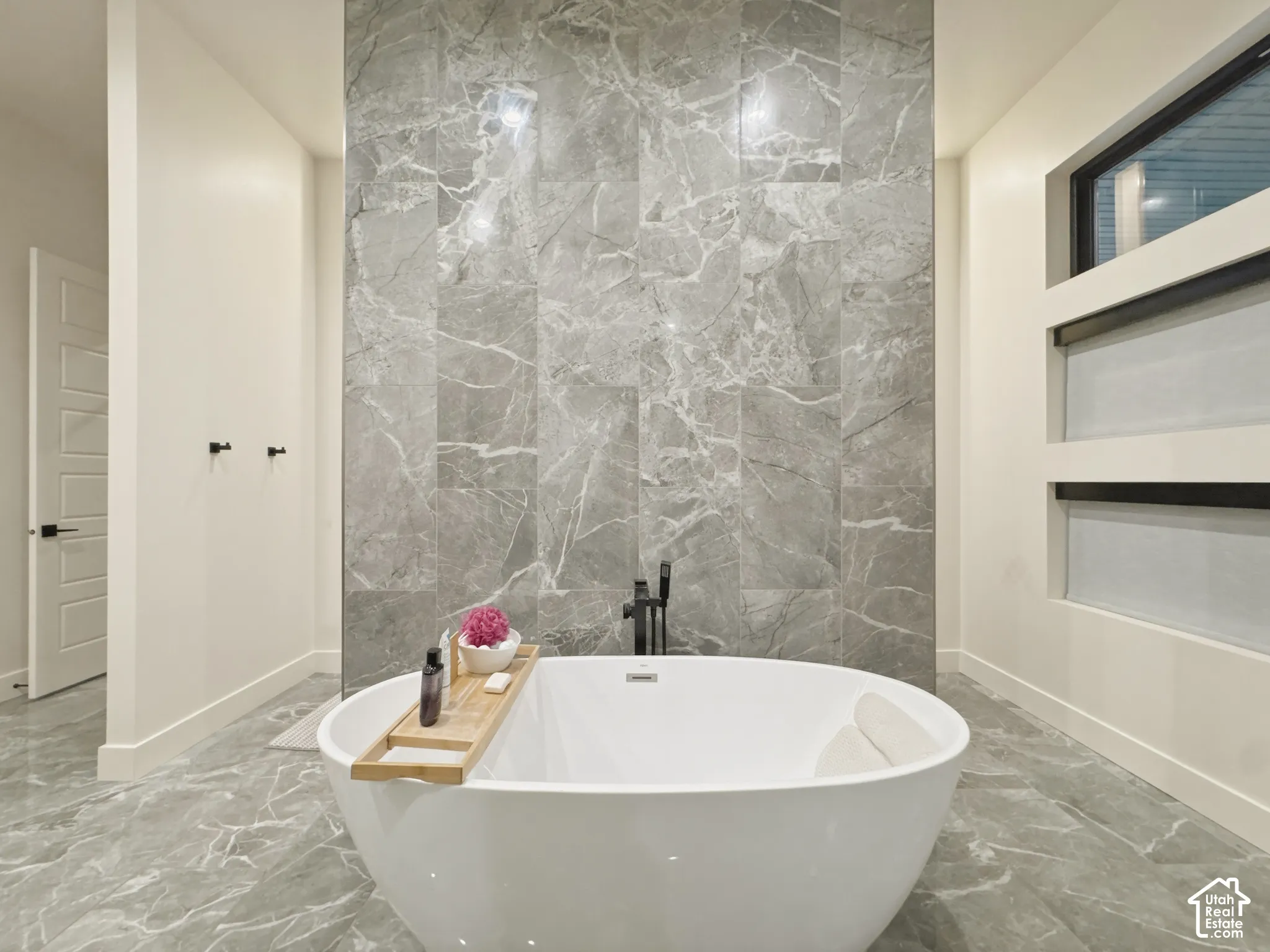Freestanding soaking tub with matte black fixtures, marble-patterned tile flooring, full-height marble-patterned accent wall, and built-in privacy windows