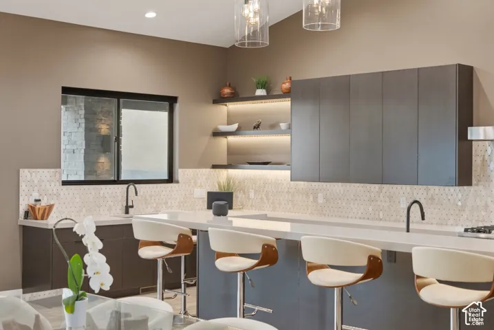 Kitchen featuring a light hexagonal tile backsplash, dark flat-panel cabinetry, light countertops, and integrated open shelving with under-cabinet lighting