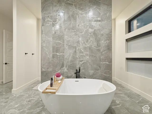 Freestanding soaking tub with matte black fixtures, marble-patterned tile flooring, full-height marble-patterned accent wall, and built-in privacy windows
