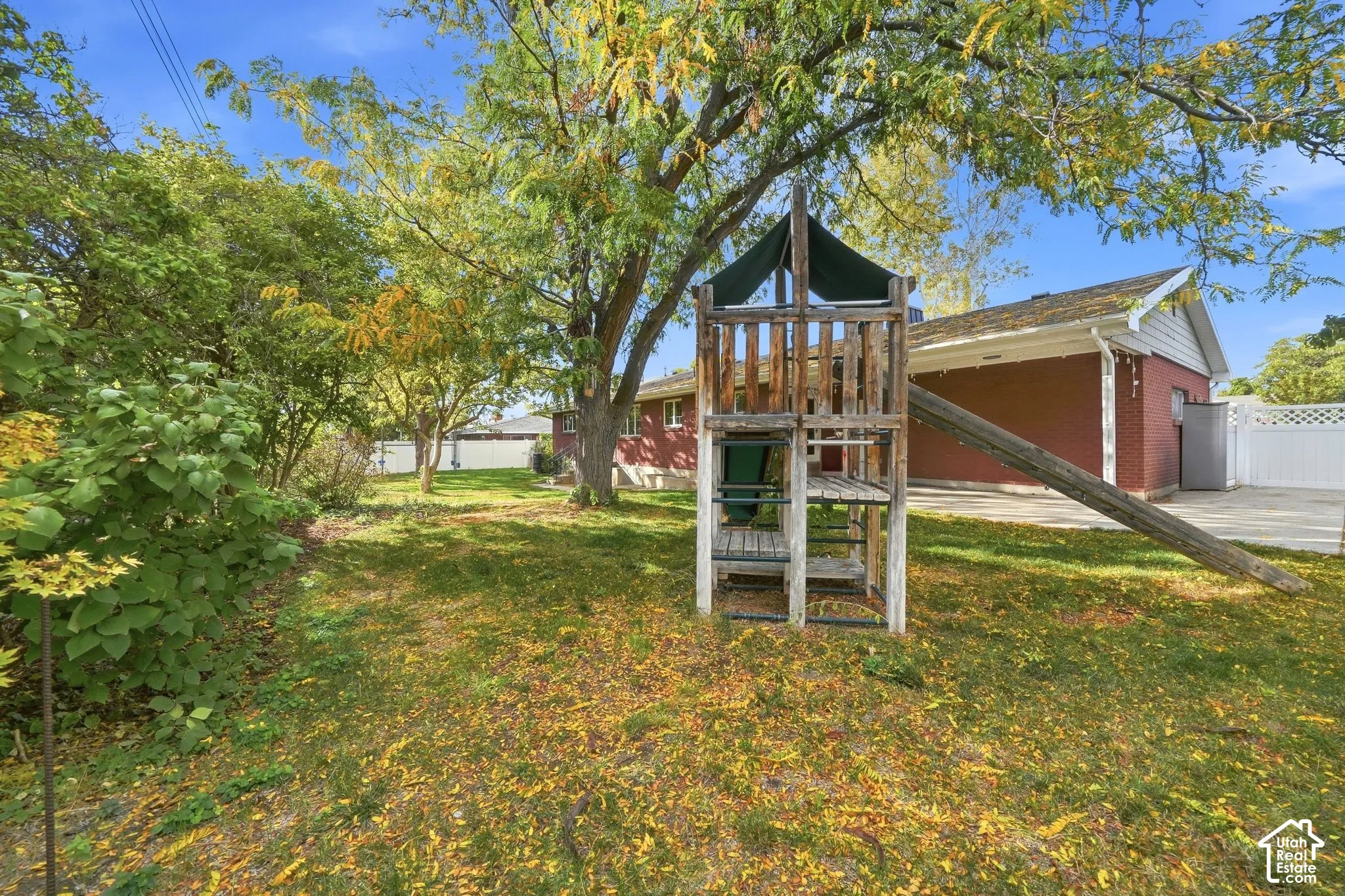 Wooden play fort with slide under the trees, patio and home beyond.