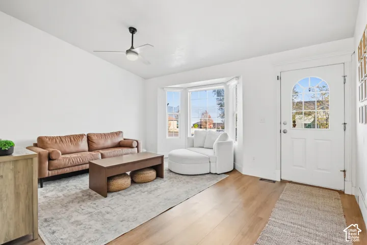 Wood-finish flooring, bay window, white entry door with arched glass, ceiling fan, and white walls