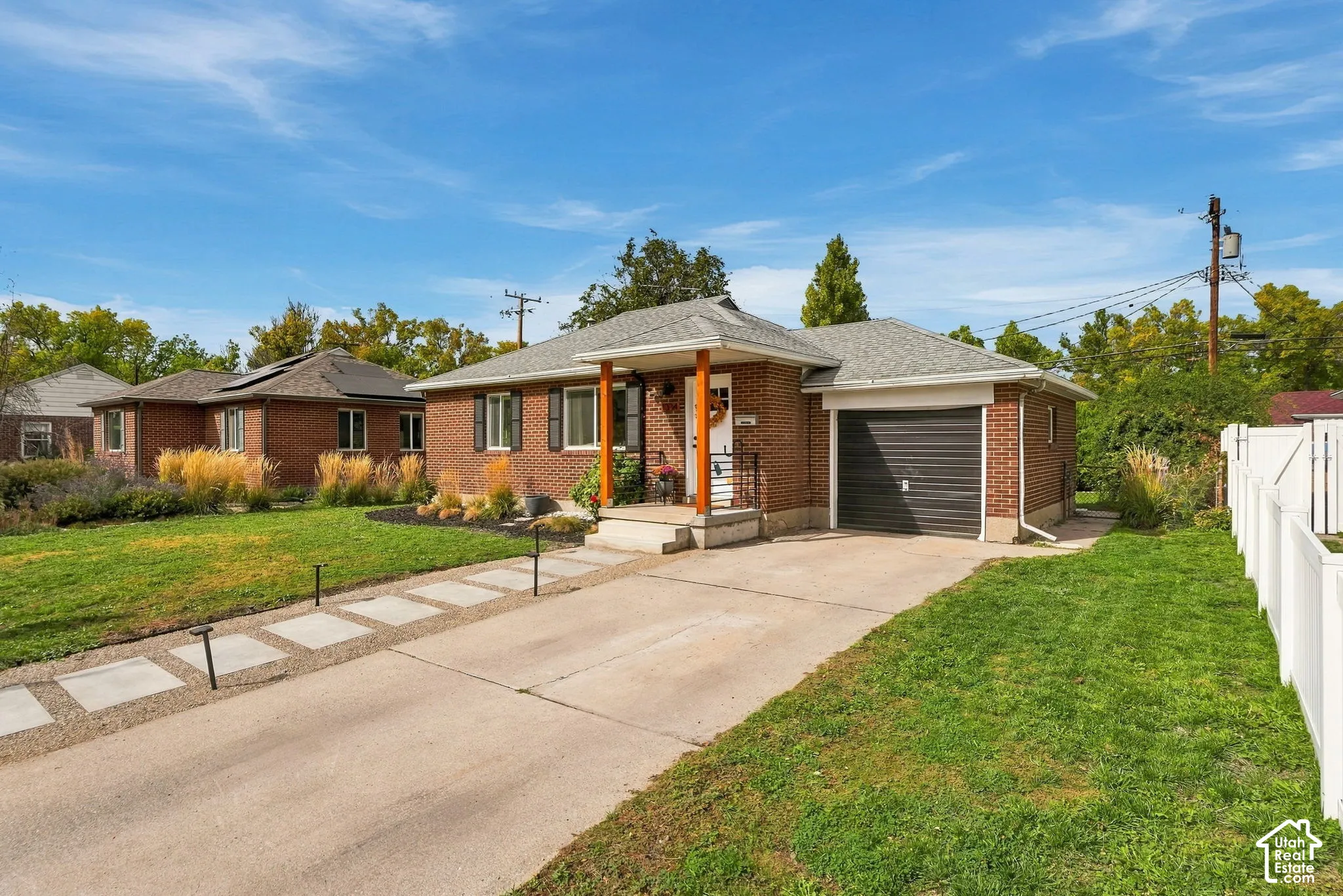 Brick exterior with a gabled roof, attached garage, concrete driveway, front porch, and partial white fencing